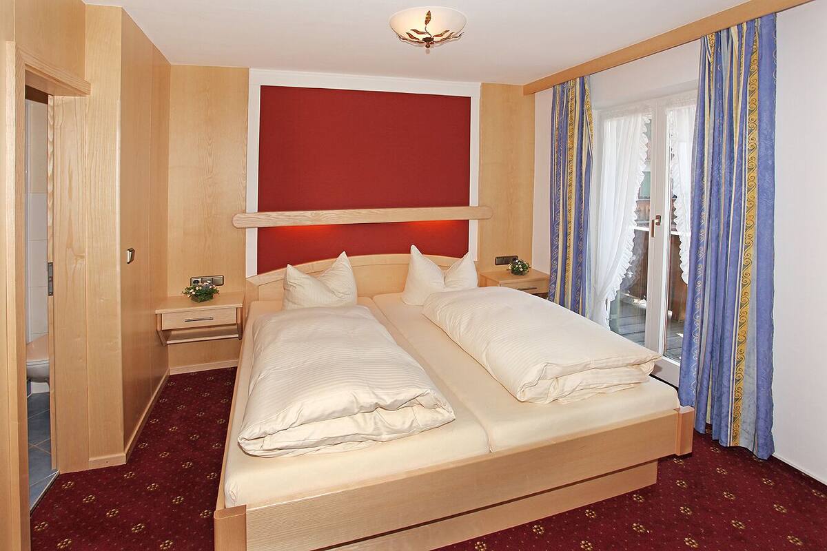 Een tweepersoonsbed met houten frame, een rode accentmuur, een nachtkastje met lamp en een raam met gordijnen.