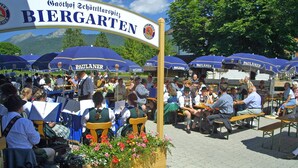 Un jardin à bière avec des tables et des chaises, entouré de parasols bleus sur lesquels on peut voir la marque « Paulaner ».