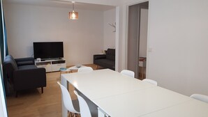 Ein modernes Wohnzimmer mit Flachbildfernseher, einer Couch, einem Esstisch mit Stühlen und einer Pendelleuchte.