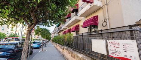 A street view with parked cars, a tree-lined sidewalk, and a building with balconies and awnings.
