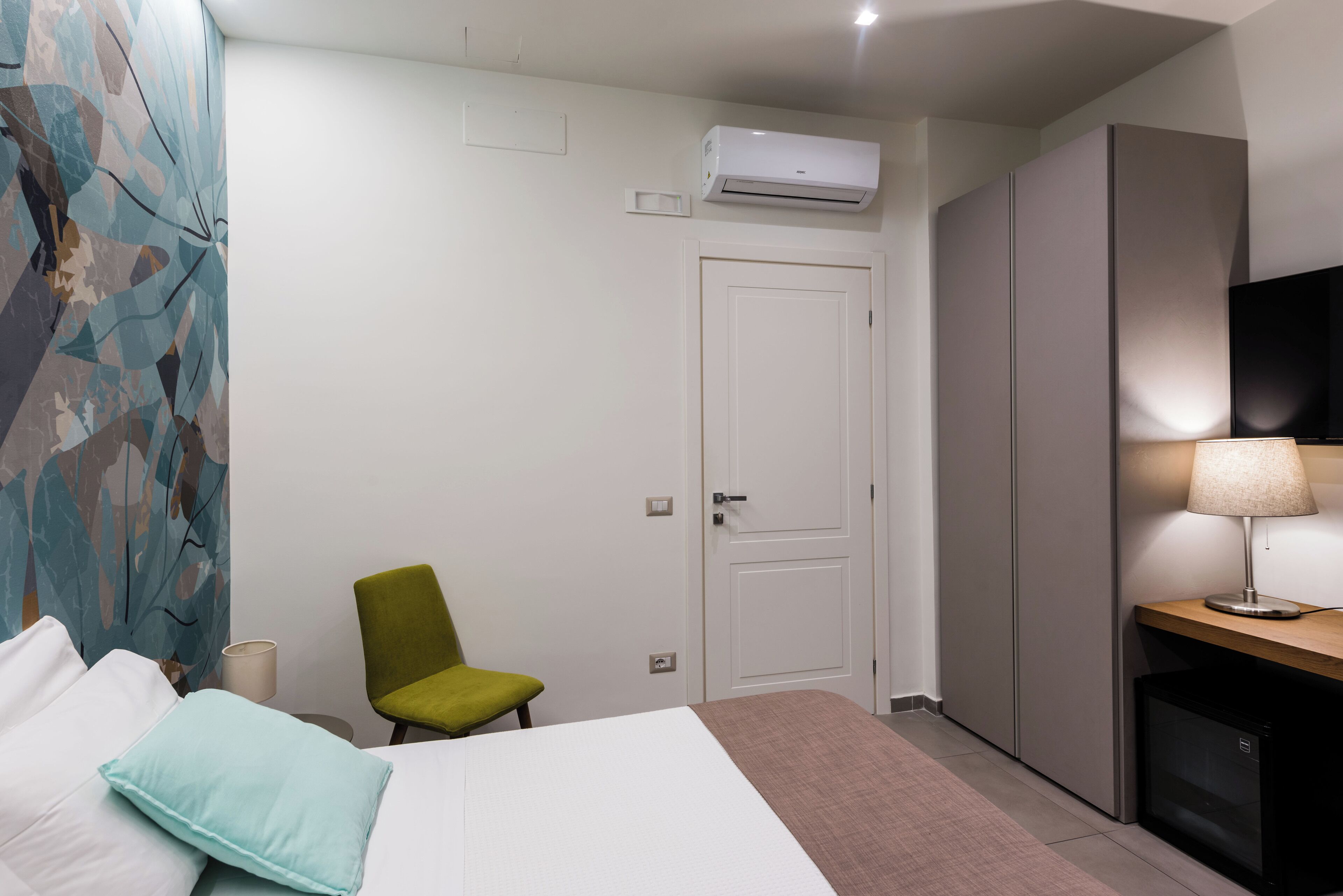 A hotel room with a bed, a green chair, a desk with a lamp, a wardrobe, and an air conditioning unit.