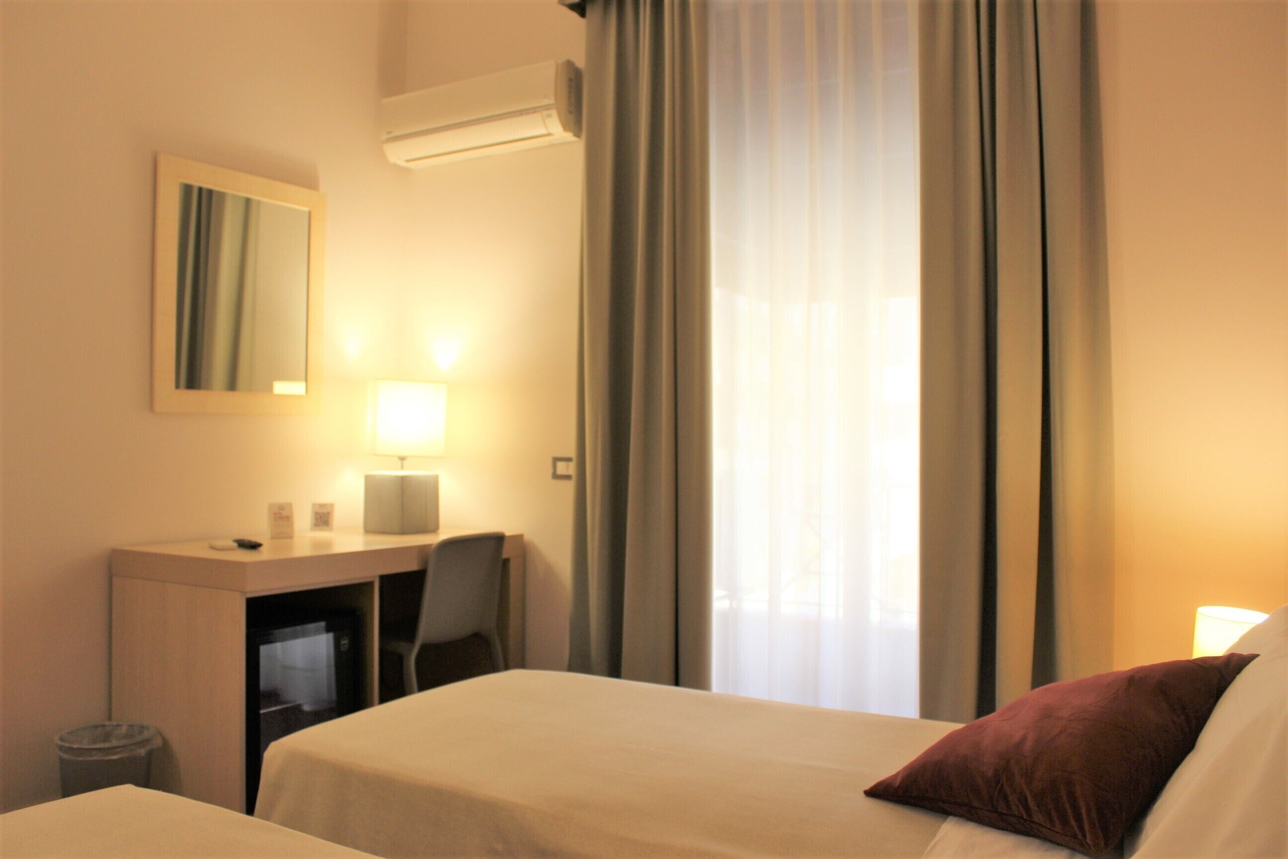 Hotelkamer met een bed, een bureau met stoel, een prullenbak, een spiegel en een raam met gordijnen.
