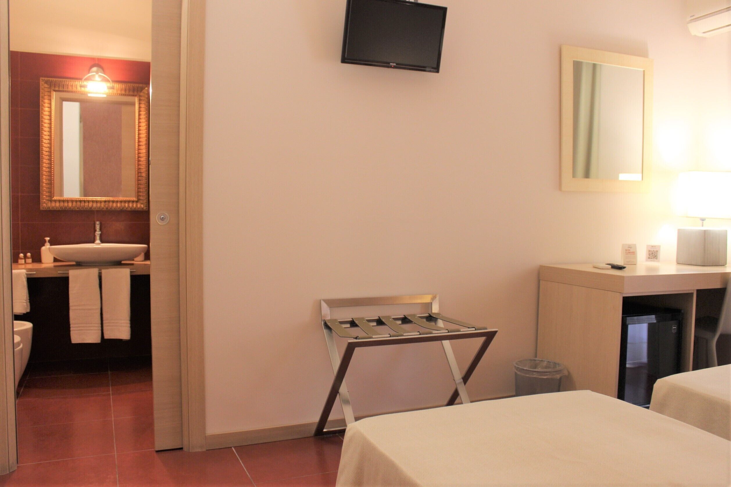 Habitación de hotel con cama, escritorio, televisión, espejo y baño con lavamanos y toallas.