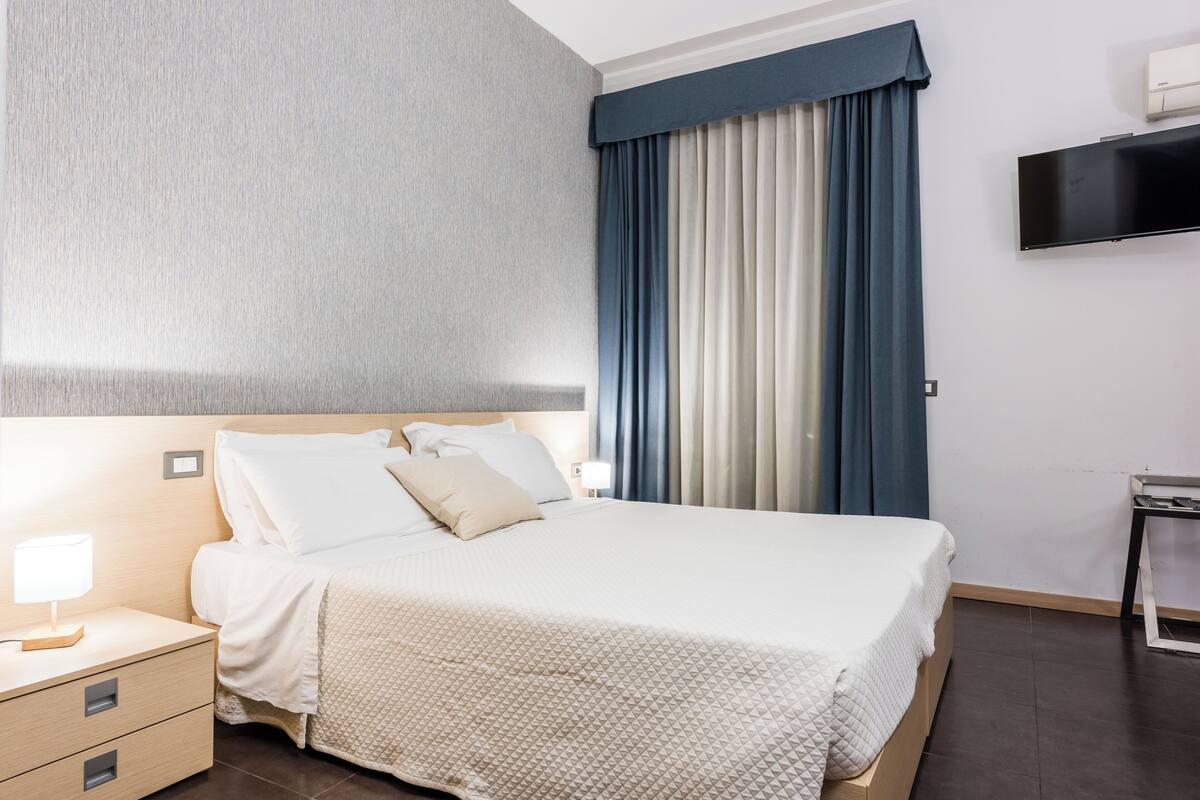 A hotel room with a bed, bedside tables, a flat-screen TV, and a large window with curtains.