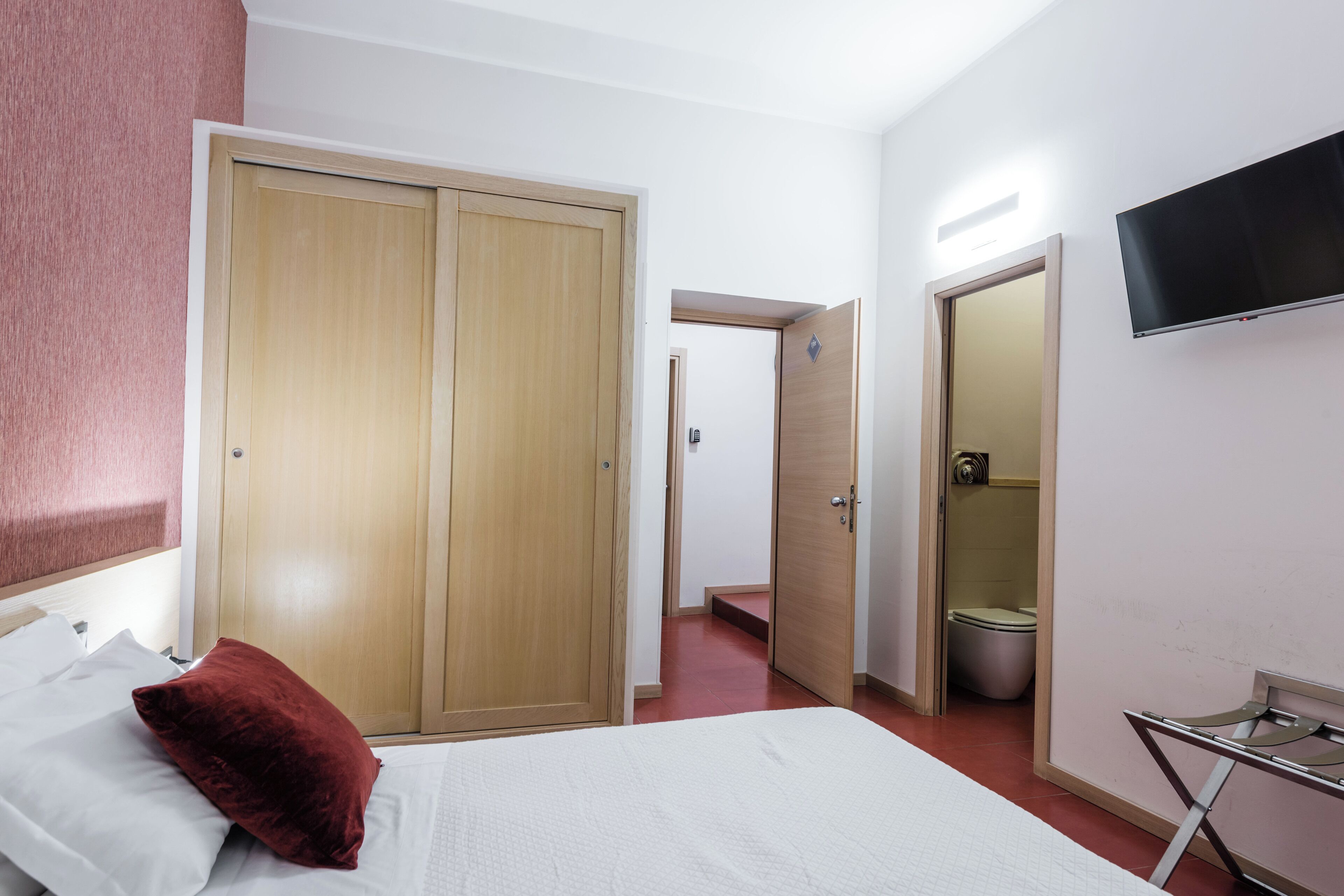 A hotel room with a bed, a red pillow, a wardrobe, a television, and a bathroom.