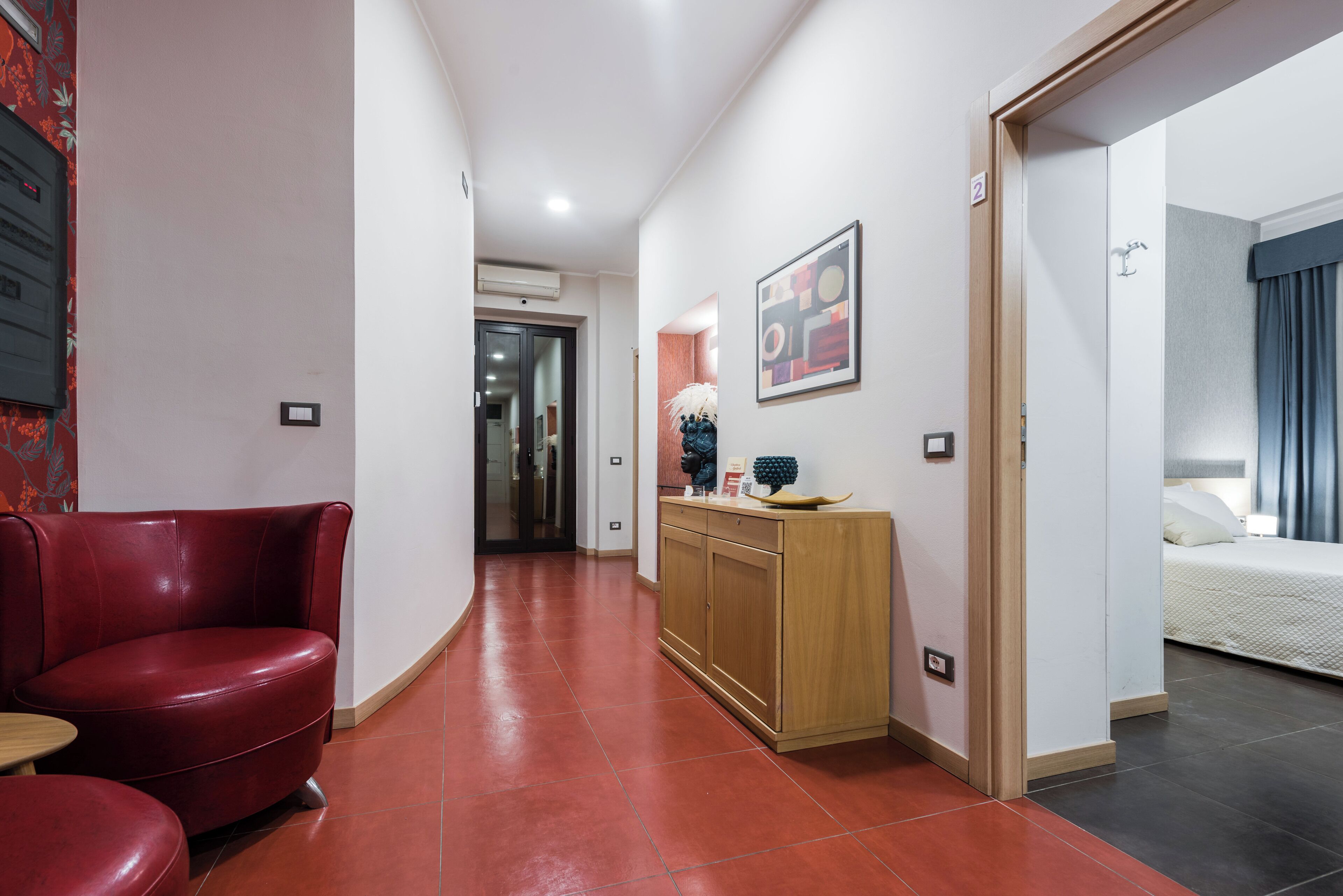 A hotel corridor with a red leather chair, a wooden reception desk, and a bed in the adjacent room.