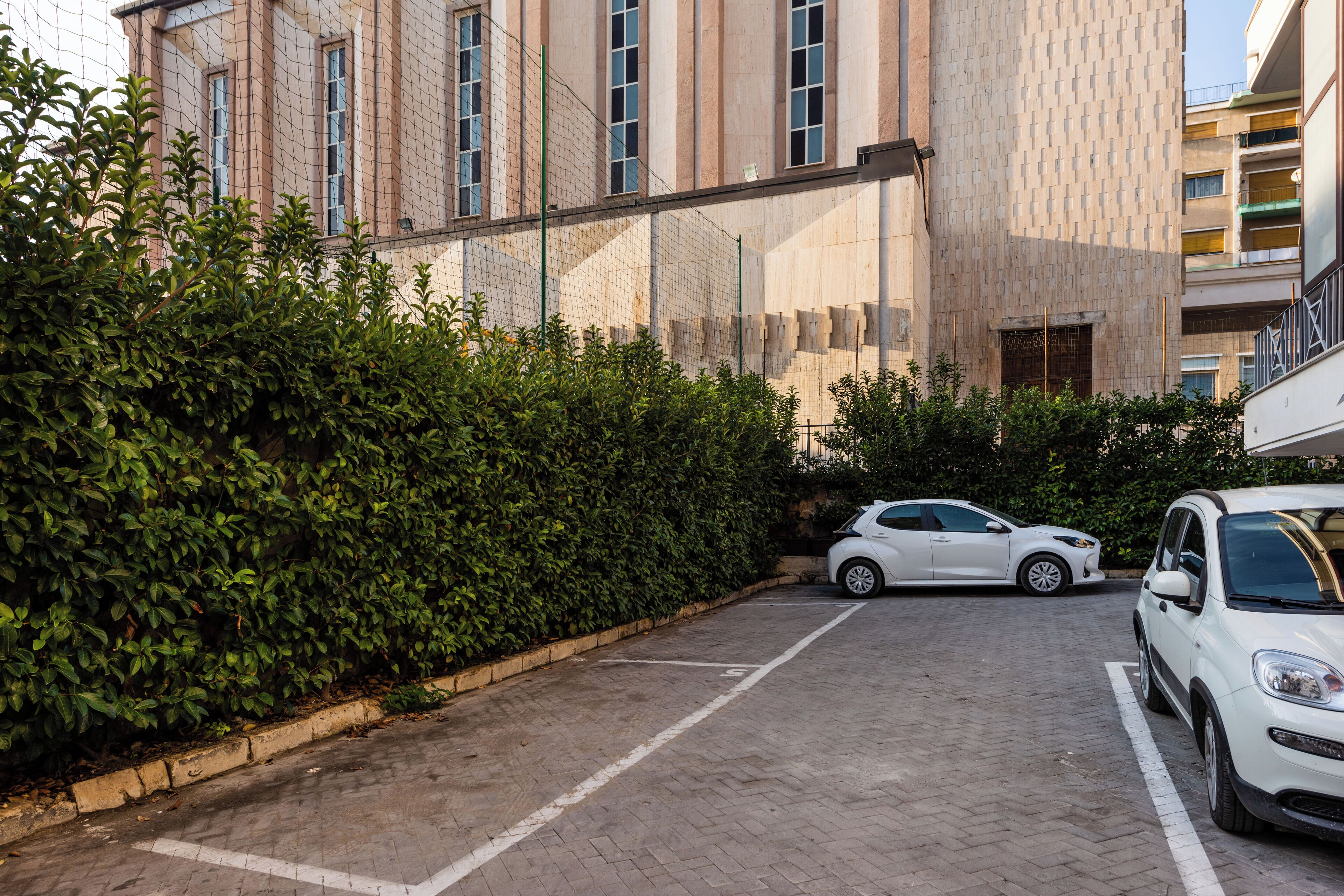 A parking area with two white cars, a hedge, and a building with large windows and a textured facade.