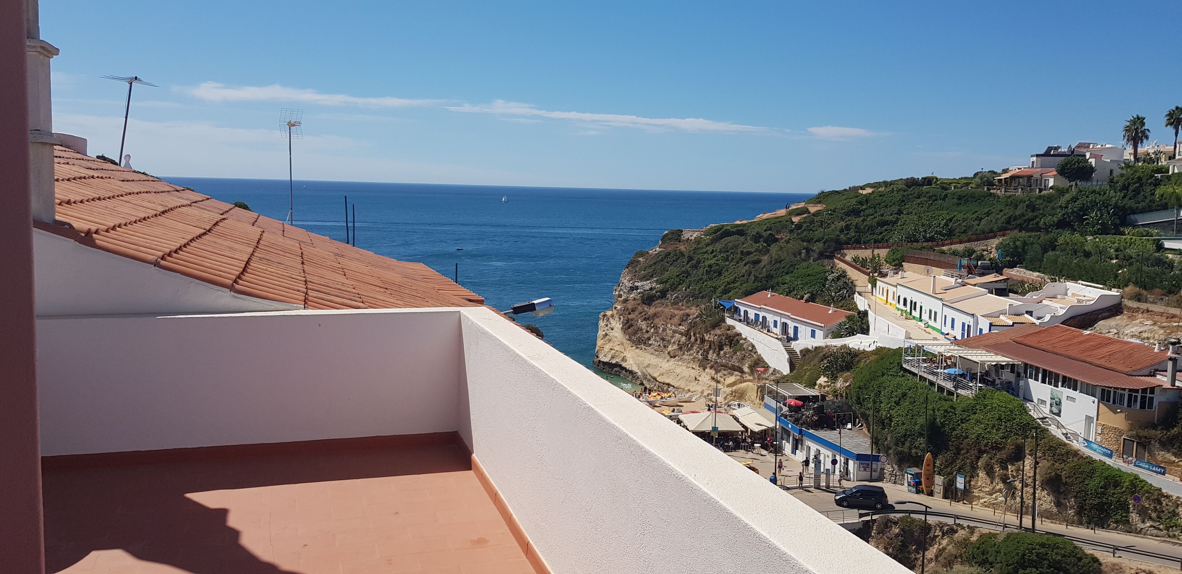 A coastal view with a balcony overlooking the sea, buildings with red roofs, and a clear sky.