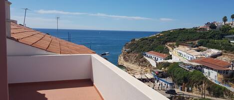 A coastal view with a balcony overlooking the sea, buildings with red roofs, and a clear sky.
