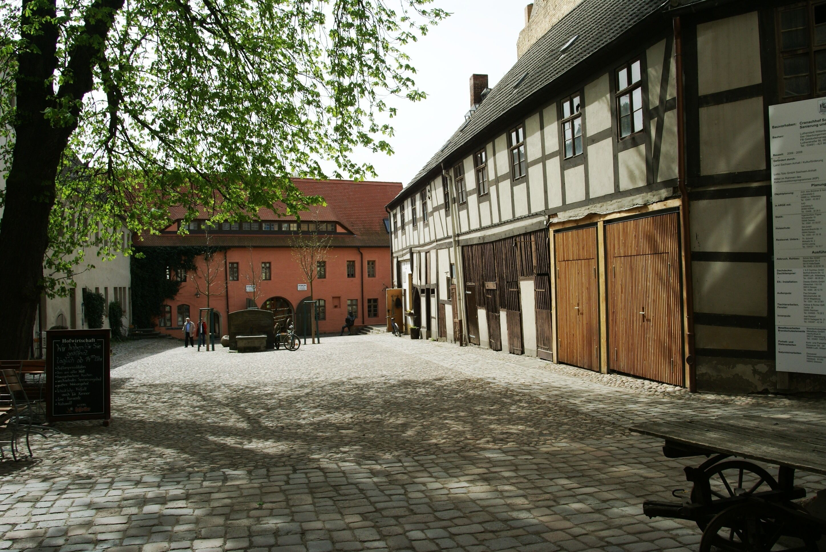 A cobblestone courtyard with a traditional half-timbered building, a red brick building, and a signboard.