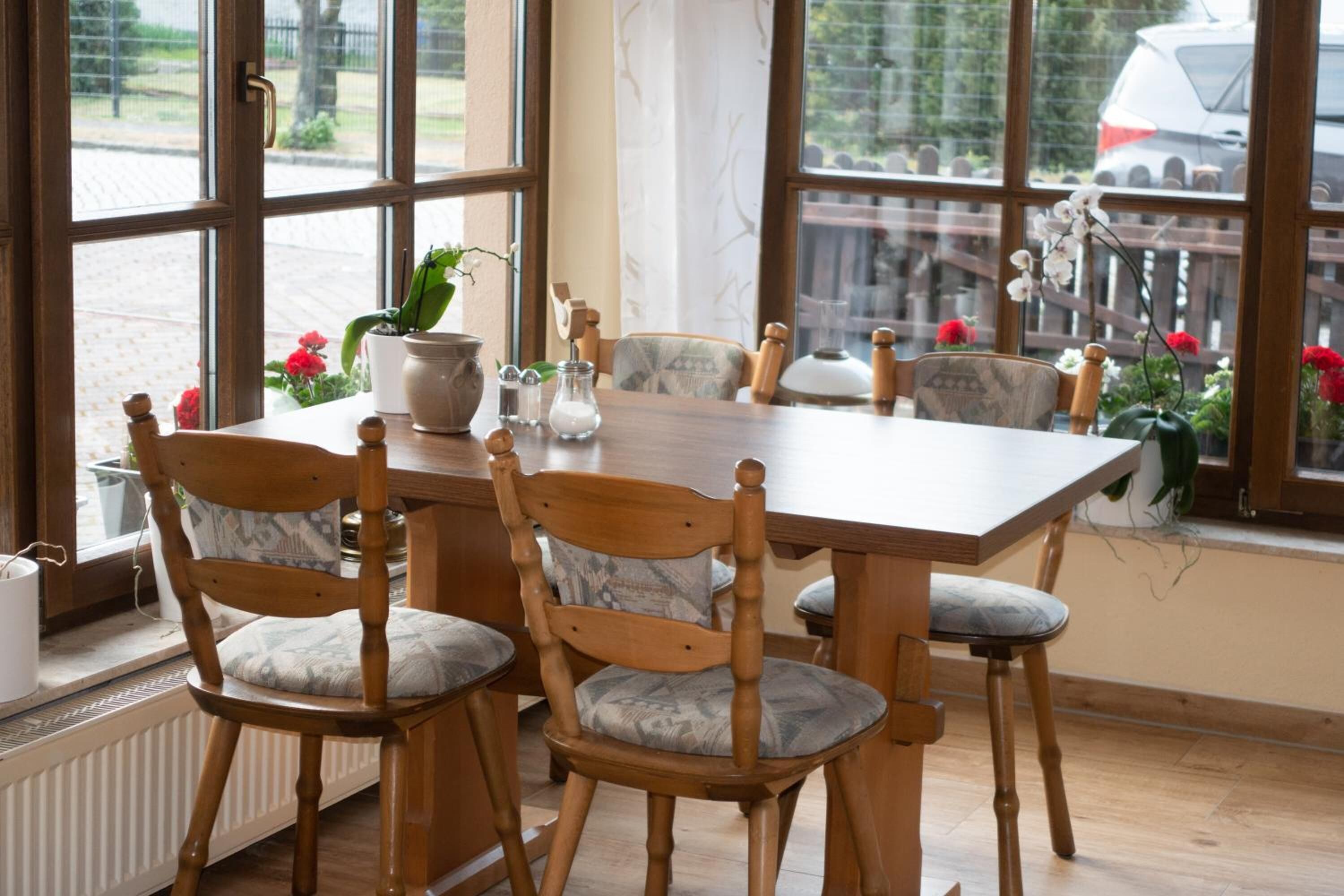 A dining area with a wooden table and chairs, potted plants, and a view through large windows.