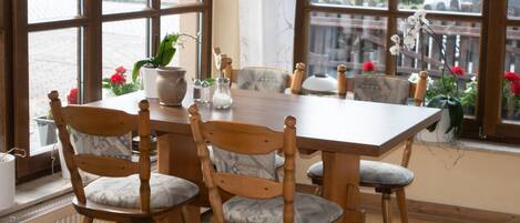 A dining area with a wooden table and chairs, potted plants, and a view through large windows.