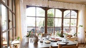 Ein Essbereich mit einem Holztisch, gedeckt für zwei Personen, umgeben von Stühlen. Üppige Kübelpflanzen, ein Teekessel, Tassen und Teller mit Speisen sind ebenfalls vorhanden. Große Fenster mit Vorhängen sorgen für natürliches Licht und einen Ausblick ins Freie.