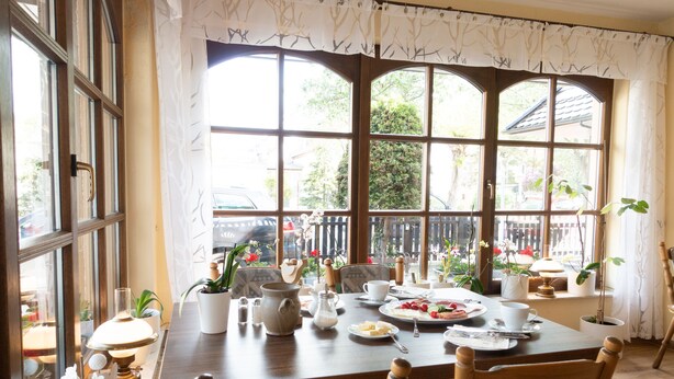 Un coin repas avec une table en bois dressée pour deux personnes, entourée de chaises. On y trouve des plantes en pot, une théière, des tasses et des assiettes remplies de nourriture. De grandes fenêtres, ornées de rideaux, laissent entrer la lumière naturelle et offrent une vue sur l’extérieur.