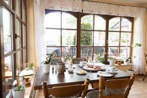 Un coin repas avec une table en bois dressée pour deux personnes, entourée de chaises. On y trouve des plantes en pot, une théière, des tasses et des assiettes remplies de nourriture. De grandes fenêtres, ornées de rideaux, laissent entrer la lumière naturelle et offrent une vue sur l’extérieur.