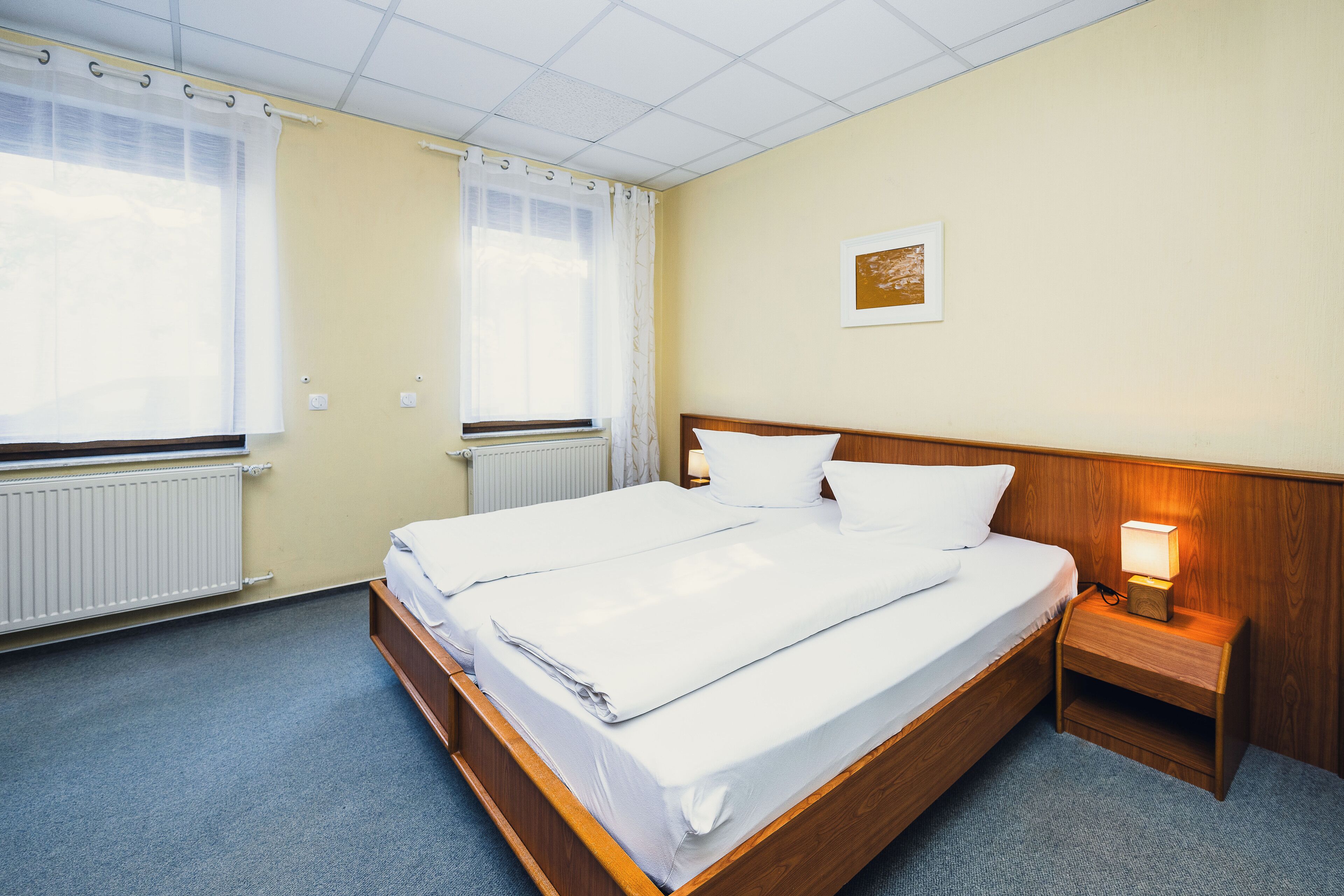 A double bed with white linens, a wooden headboard, a bedside table with a lamp, and a window with sheer curtains.