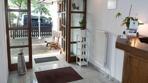 Un espace d’accueil avec un bureau en bois, une étagère blanche, une plante en pot et une vue sur l’extérieur grâce à une porte vitrée.