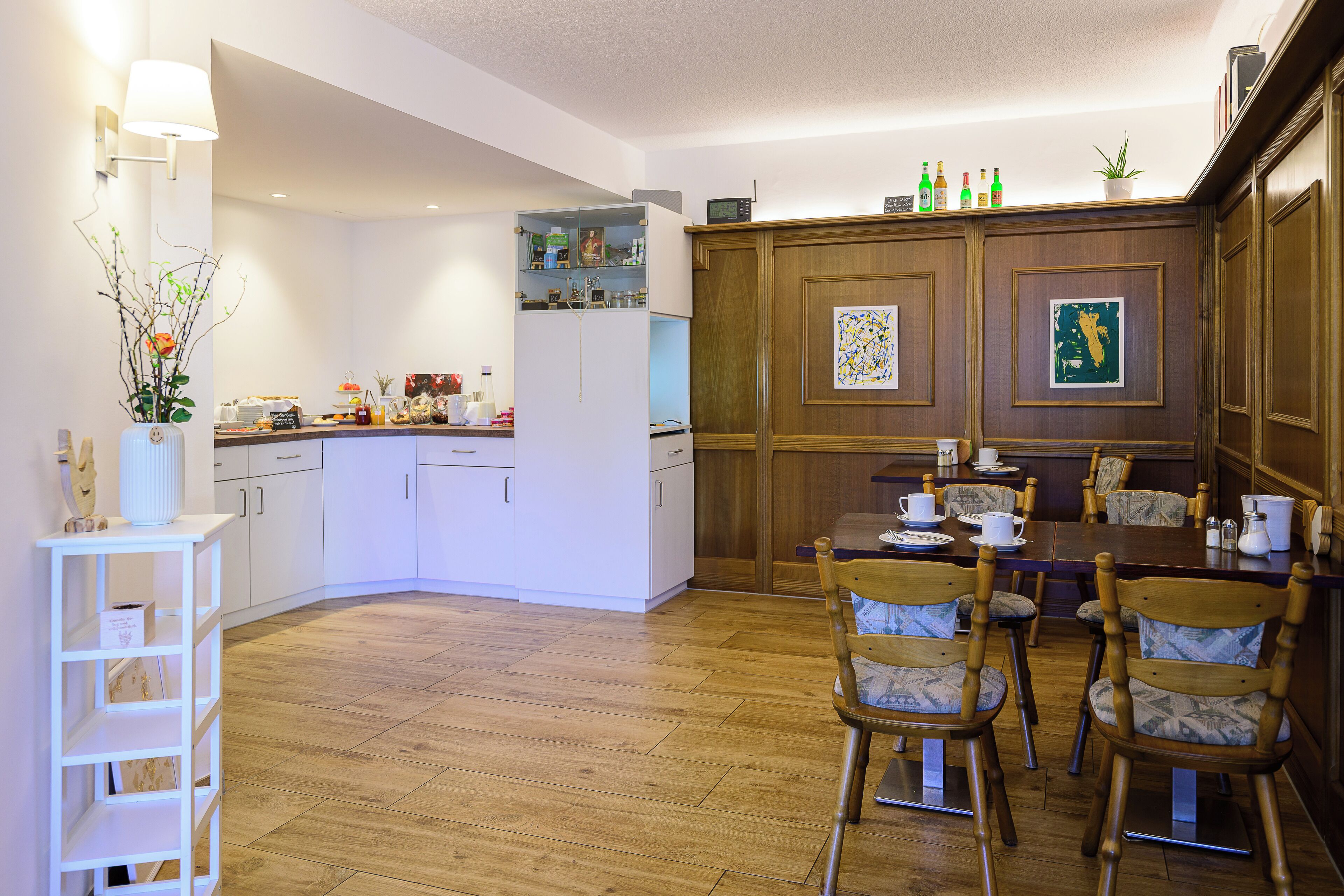 A dining area with wooden chairs and a table, a kitchen with white cabinets, and a wooden cabinet with framed artwork.