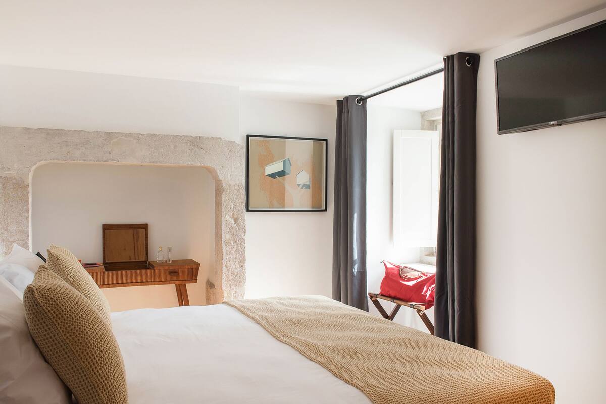 Una habitación de hotel con una cama, una mesita de noche de madera, un bolso rojo, un televisor y un cuadro en la pared.