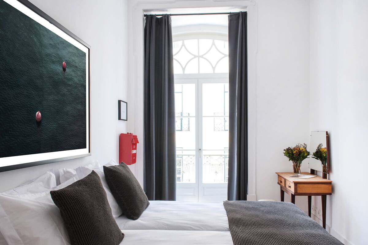 Una habitación de hotel con cama, almohadas, una mesita de noche con un florero y un extintor rojo, además de un ventanal con cortinas.