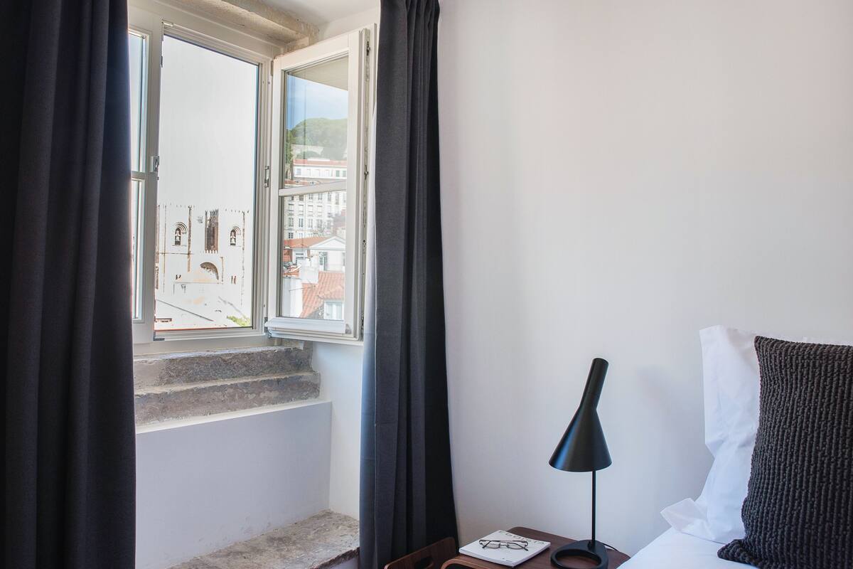 Una habitación de hotel con cama, mesita de noche, lámpara y ventana con cortinas.