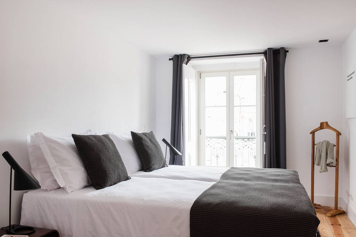 Un dormitorio con cama, una mesita de noche, una lámpara colgante, una ventana con cortinas y suelo de madera.
