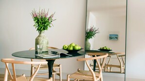 Un coin repas avec une table ronde en verre, deux chaises en bois, un vase avec des fleurs, un saladier de pommes et un grand miroir qui reflète la pièce.