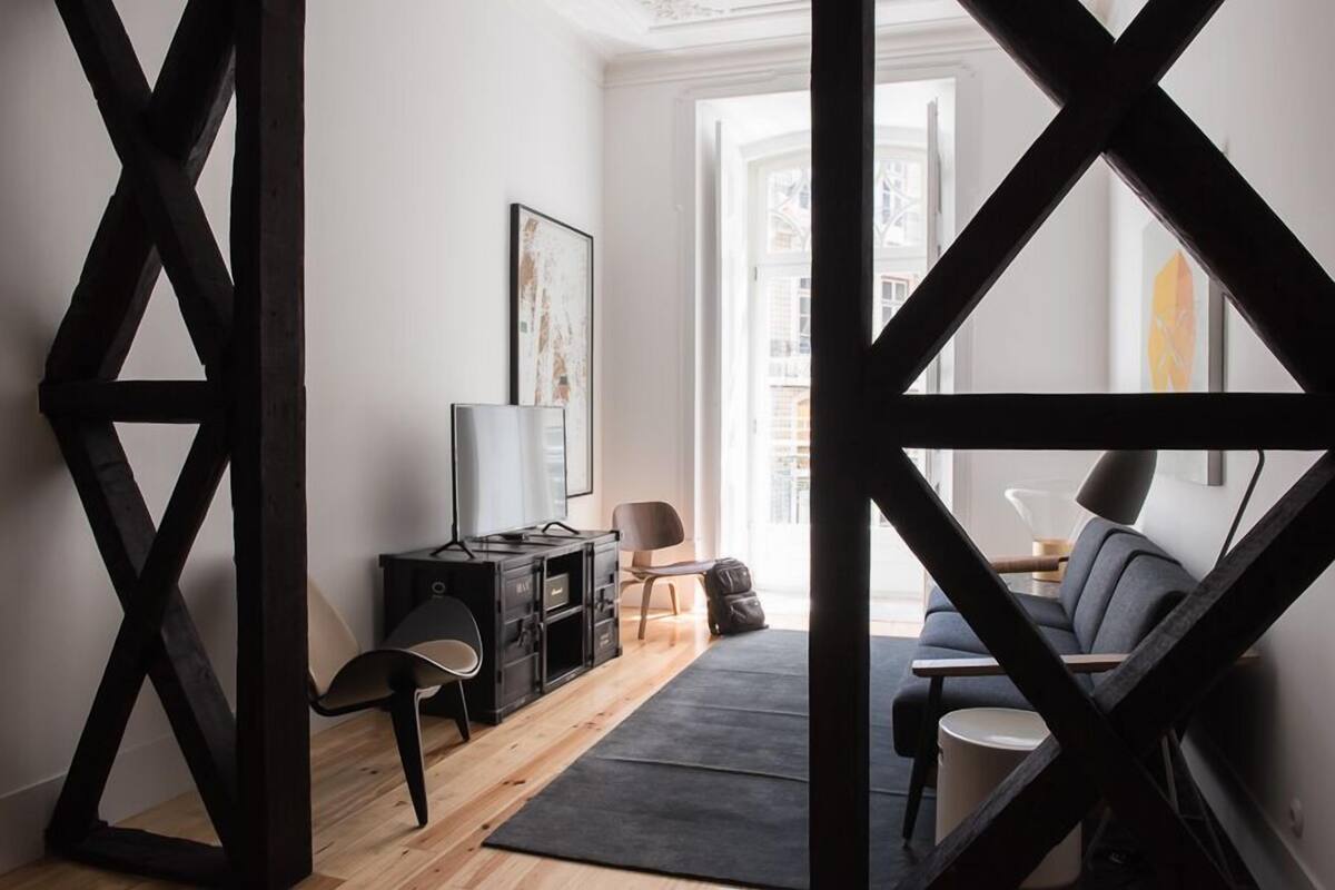 Una sala moderna con una estructura metálica negra en forma de X, un sofá gris, piso de madera y un televisor sobre un mueble negro.