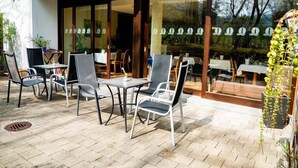 Un patio al aire libre con mesas y sillas, una zona cubierta y vistas al interior a través de amplios ventanales.