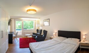 A hotel room with a bed, bedside tables, a desk, a chair, a red rug, a window, and a ceiling light. - Hotel Quellenhof Bad Urach (Bad Urach)