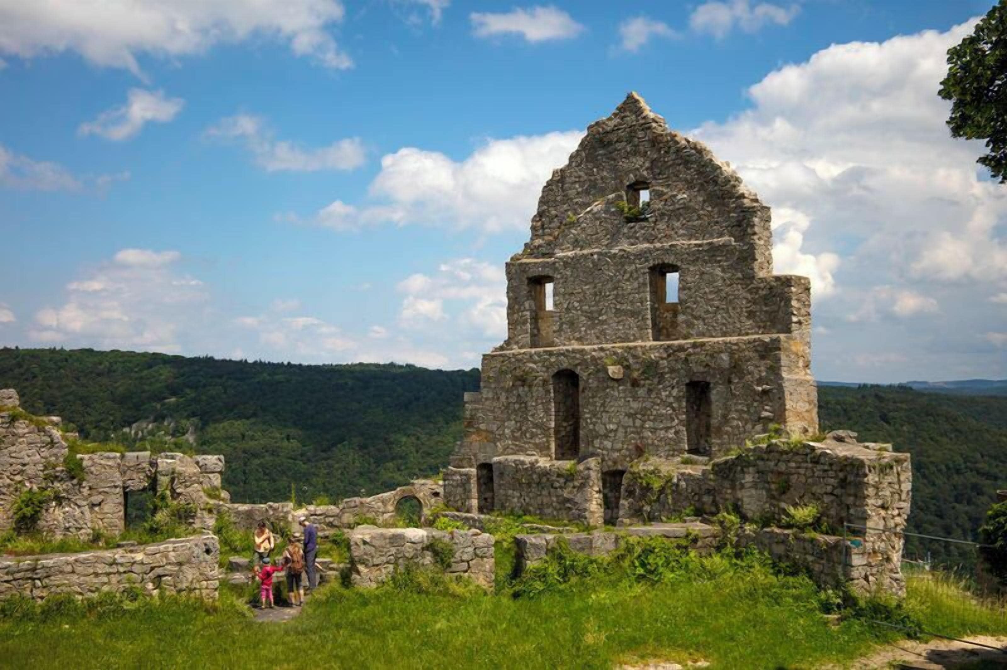 Une ancienne structure en pierre, dotée de plusieurs arches et d’un toit triangulaire, entourée d’une végétation luxuriante et avec des collines en arrière-plan.
