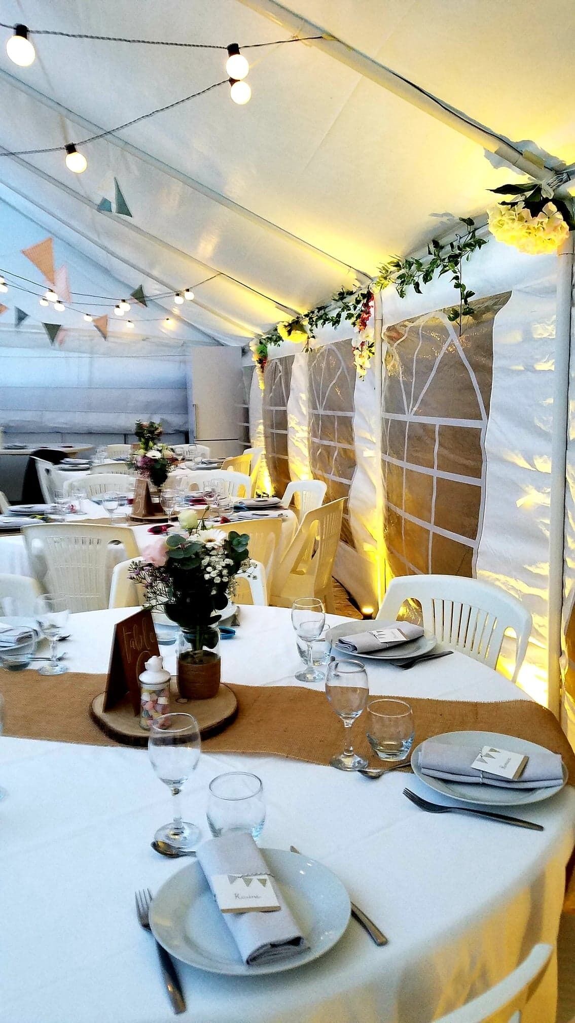 Een overdekte eethoek met gedekte tafels, lichtjes en bloemversiering.