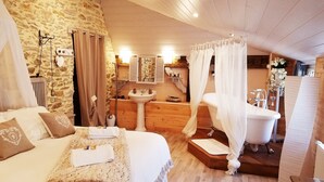 Une chambre confortable avec un mur en pierre, un lit à baldaquin, une commode en bois, un lavabo et une baignoire.