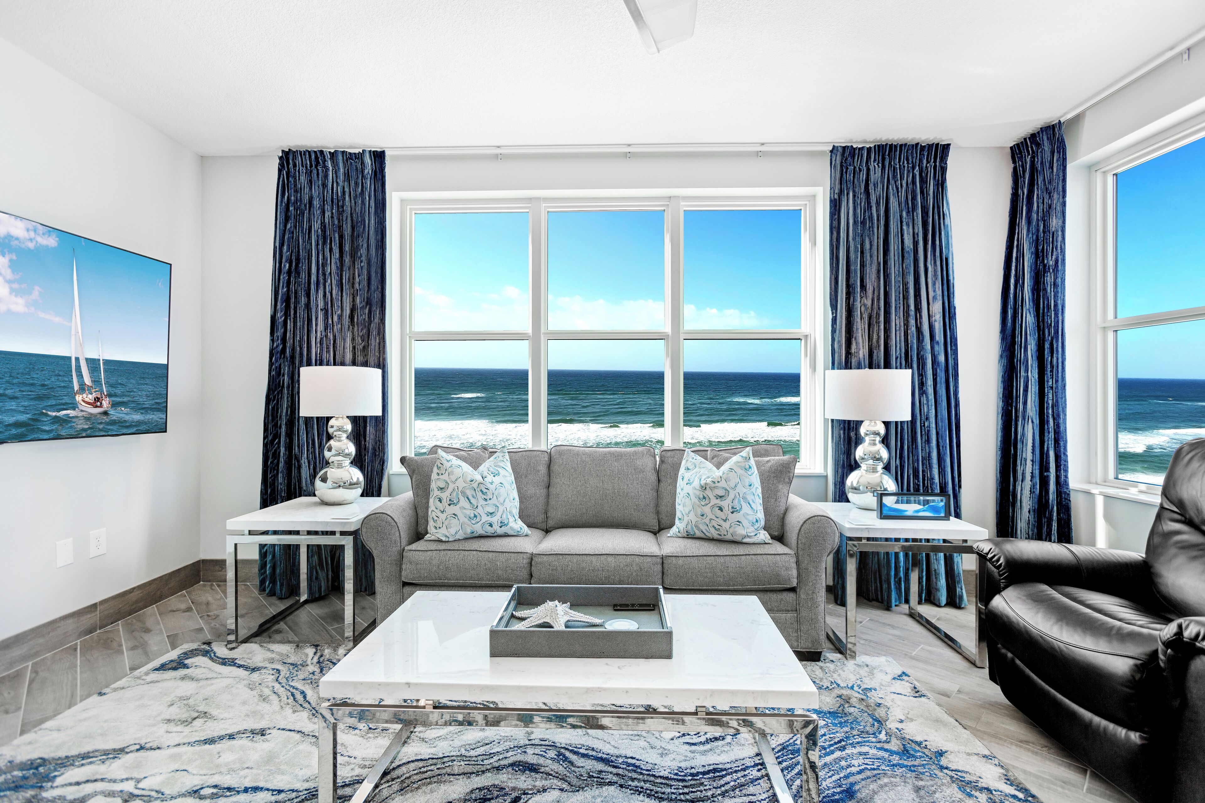A living room with a view of the ocean, a gray sofa with patterned pillows, a black leather chair, and a coffee table with decorative items.