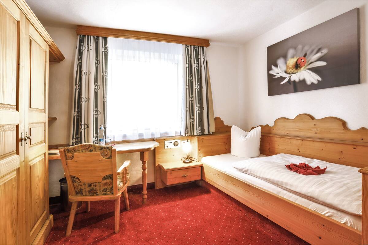 A hotel room with a wooden bed, a desk with a chair, a small bedside table, and a window with curtains.