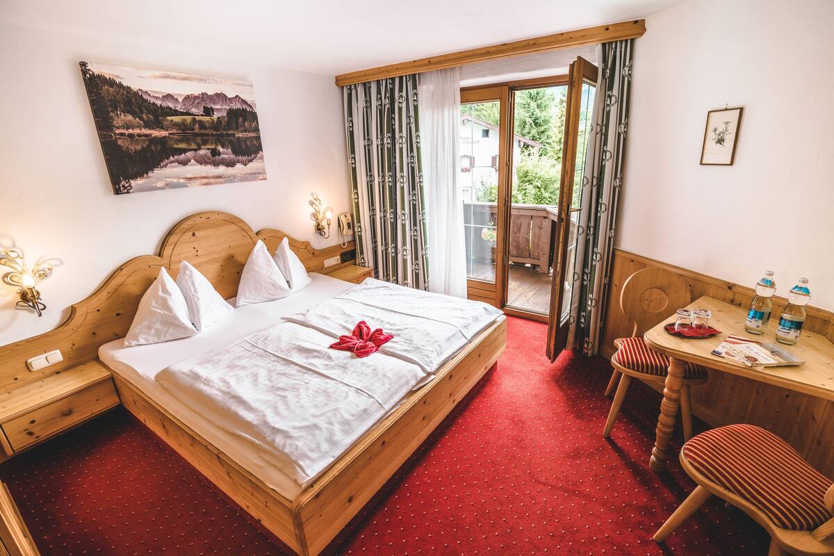 A hotel room with a wooden bed, a desk with chairs, a view of a balcony, and a painting on the wall.