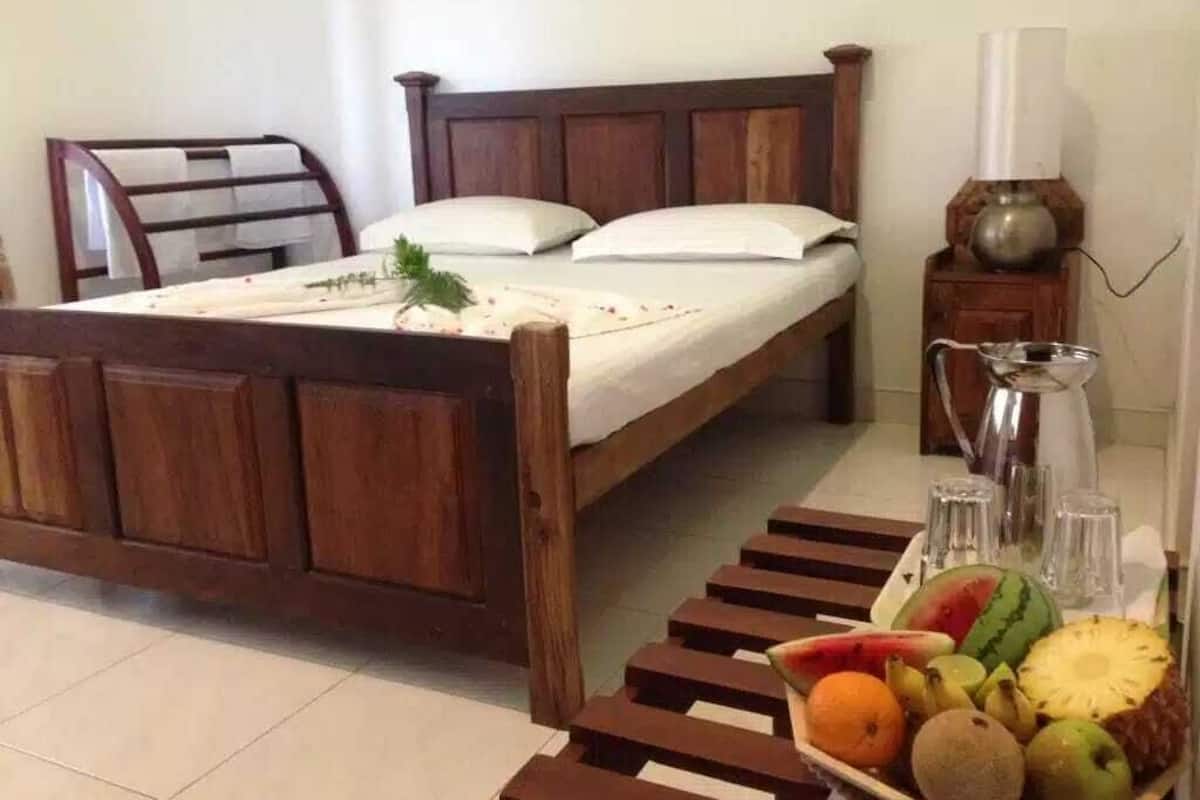 A wooden bed with a white mattress and pillows, a bedside table with a lamp, and a fruit basket on the floor.