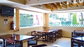 Un restaurant au décor moderne, avec des tables et des chaises en bois, un téléviseur à écran plat fixé au mur et une vue sur l’extérieur grâce à de grandes fenêtres.