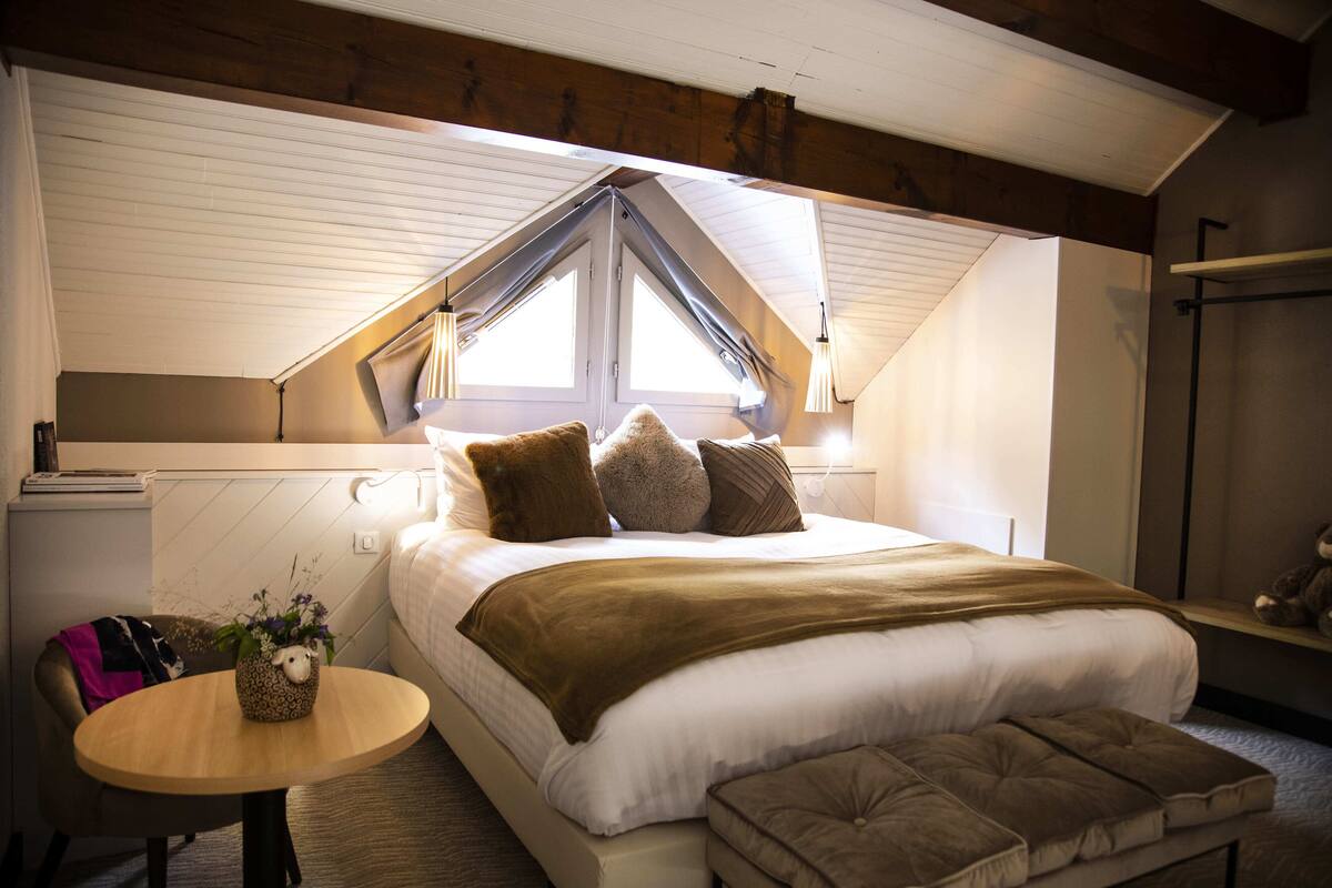 Een slaapkamer met een schuine plafond, een bed met veel kussens, een nachtkastje met een lamp, en een kleine tafel met een plant.