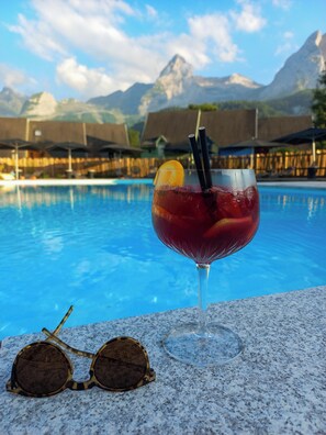 Un vaso de bebida roja con una rodaja de limón y pajitas, sobre una superficie de granito cerca de una piscina con montañas al fondo.