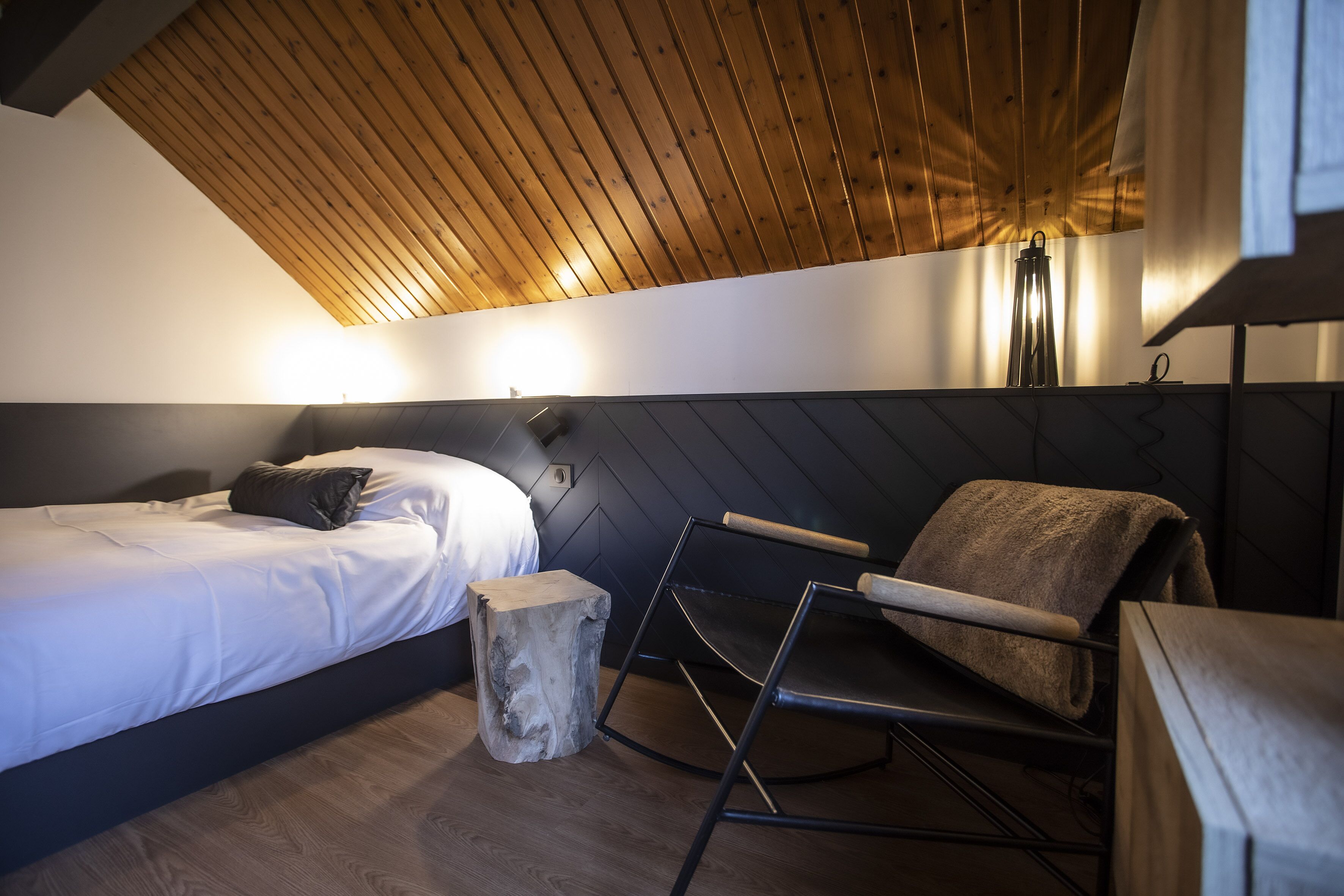 Una habitación moderna con una cama, techo de madera, un taburete pequeño y una silla plegable.