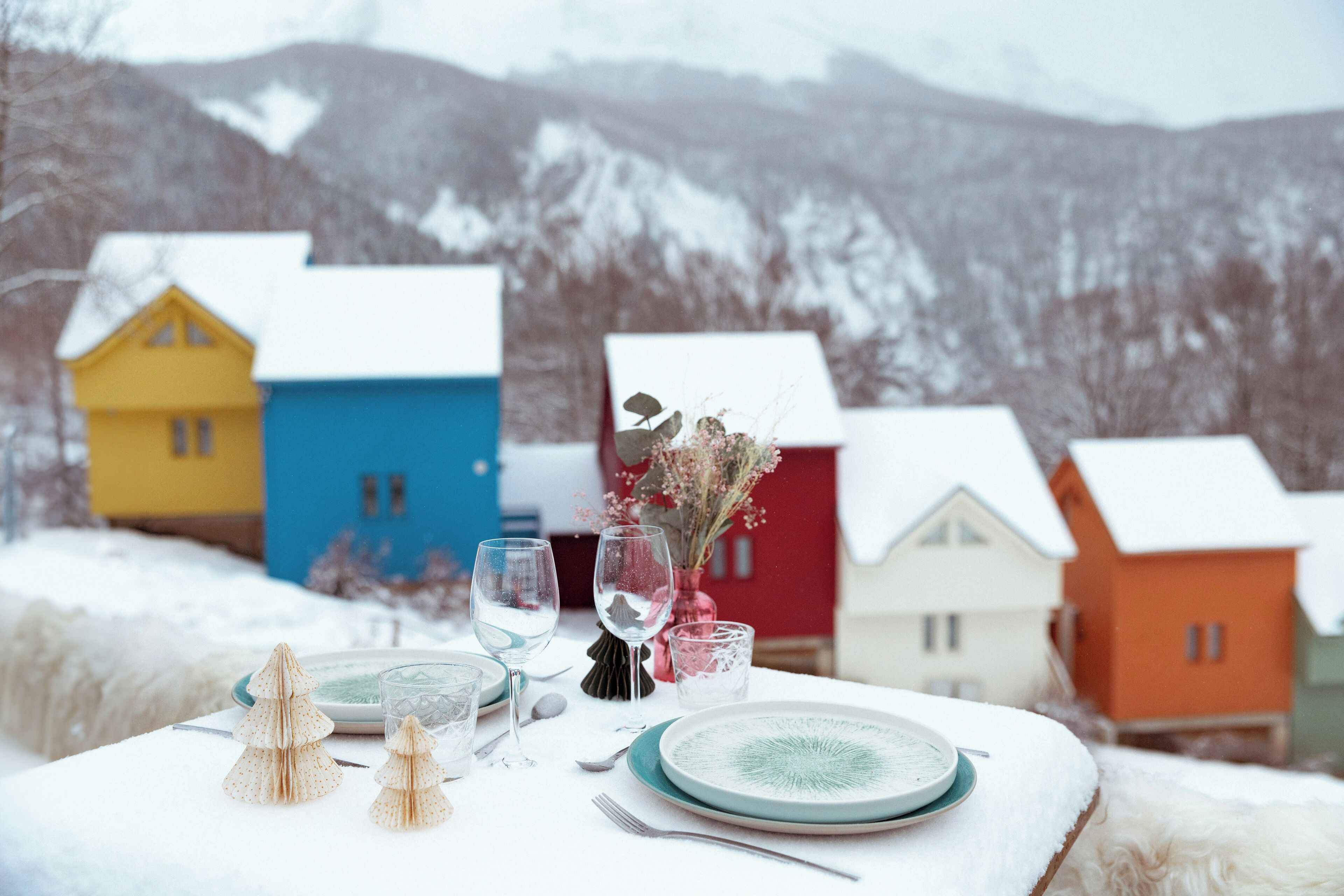 A table set with plates, glasses, and cutlery, with a snowy landscape and colorful houses in the background.