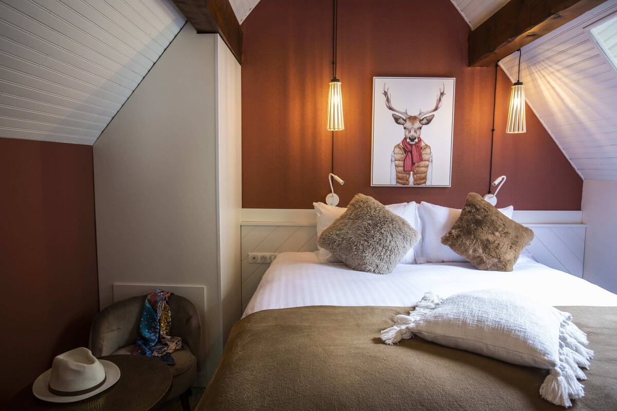 Een slaapkamer met een bed, kussens, een deken, een hoed en een schilderij van een hert op de muur.