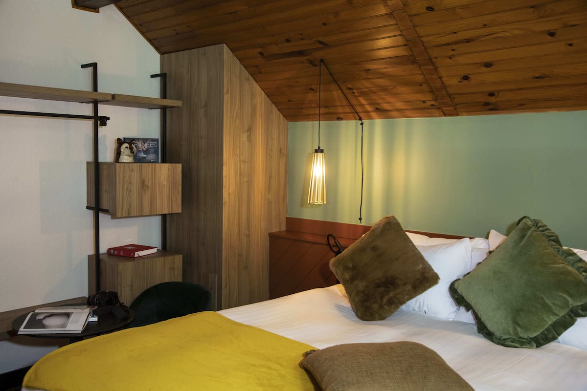 Een hotelkamer met een houten plafond, een bed met wit beddengoed, een gele plaid en een bruin kussen. Er staat een houten kastje met een rood boek en een camera erop.