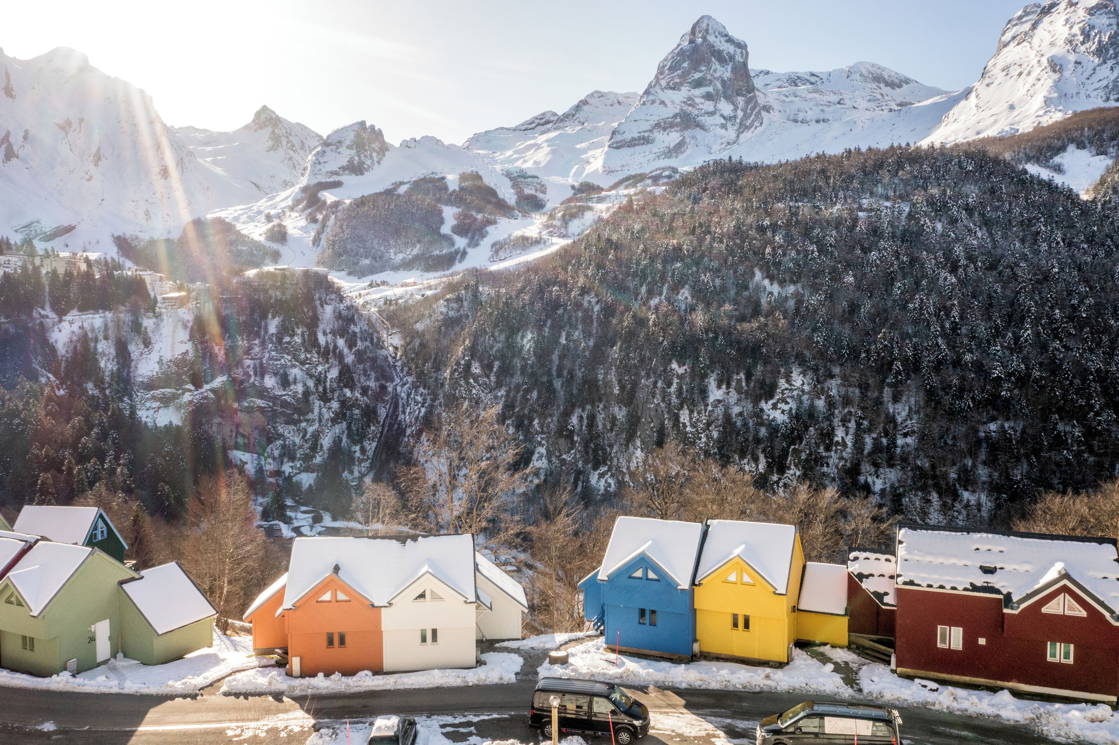 A row of colorful houses with snow-covered roofs, set against a backdrop of snow-capped mountains and a forested valley.
