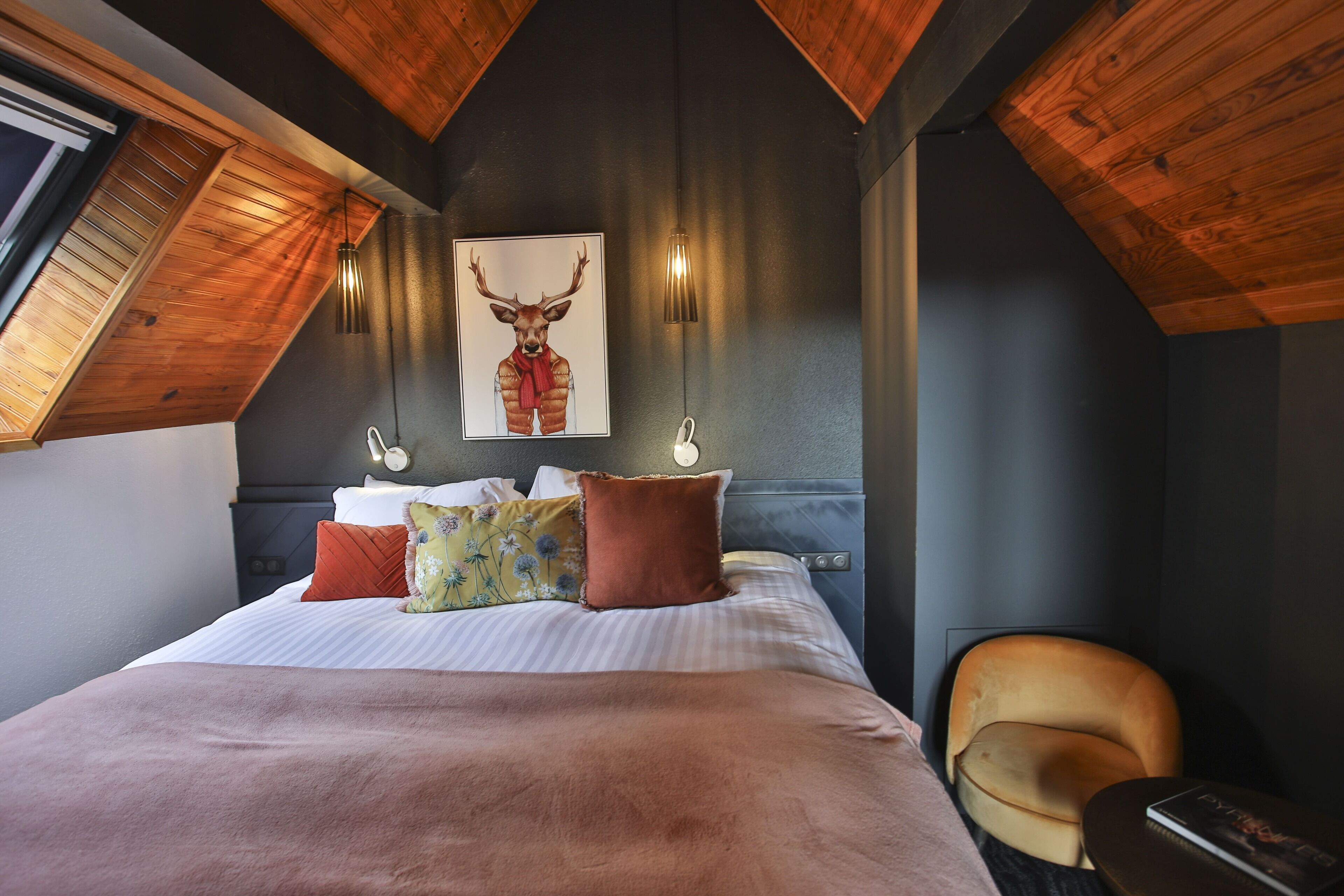 Une chambre avec un plafond en pente, un lit agrémenté de plusieurs oreillers, une chaise et une œuvre d’art représentant un cerf, accrochée au mur.