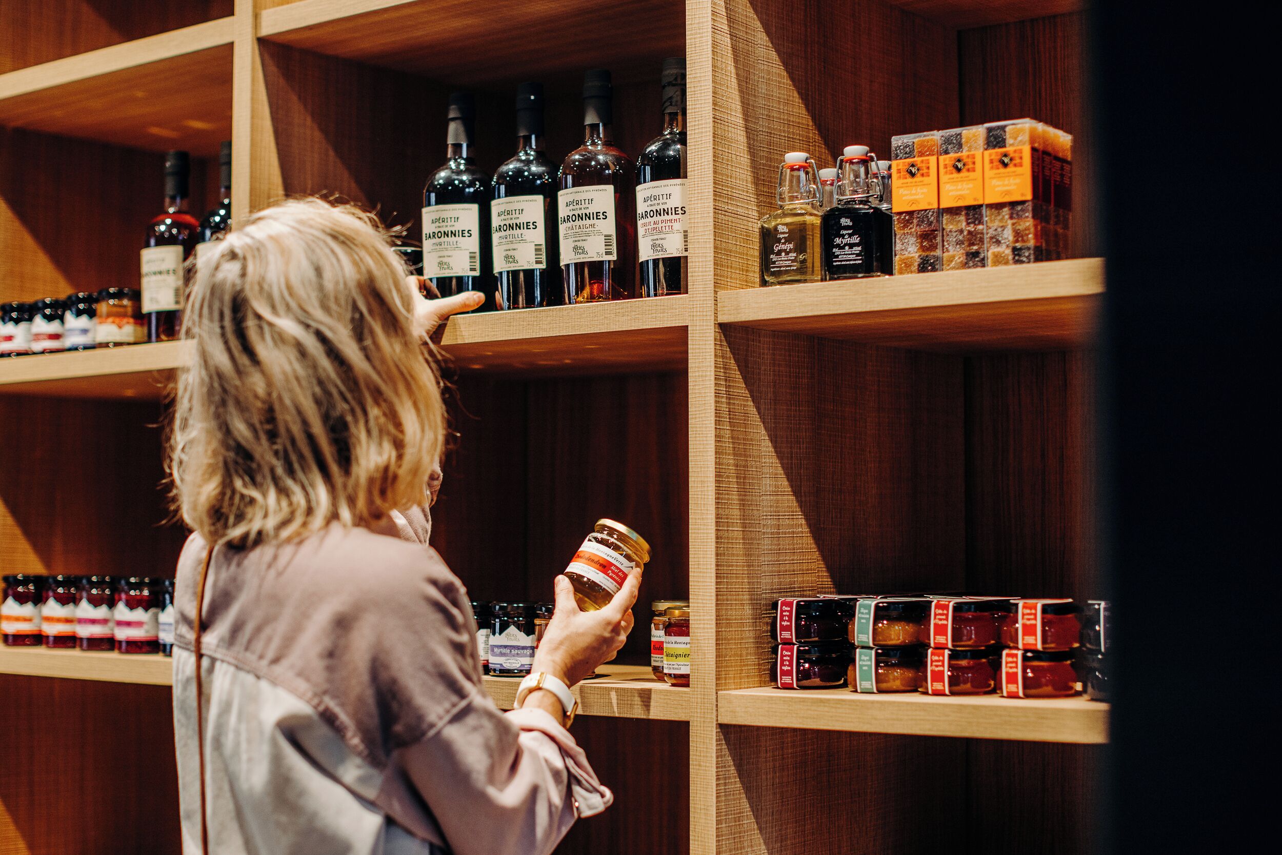 A person examining a jar of jam in a store with shelves of various bottles and jars.