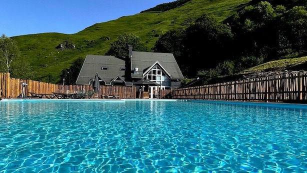 A swimming pool with clear blue water, surrounded by a wooden fence, and a house with a dark roof in the background. - ISKÖ Chalets Hotel (Eaux-Bonnes)