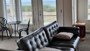 Ein Wohnzimmer mit einer schwarzen Ledersofa, einem kleinen runden Tisch und Stühlen. Große Fenster bieten Blick auf das Meer und die grüne Landschaft.