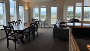 Ein geräumiger Raum mit einem großen Esstisch, mehreren Stühlen, einer Couch und einem kleinen Beistelltisch. Große Fenster bieten Blick auf das Meer und viel Grün.